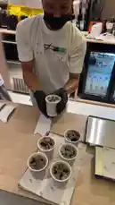 This quick look behind the counter at Sequence Coffee shows baristas preparing drinks and desserts. One barista carefully arranges six cups of coffee on a tray, while another works near a display of cakes under glass domes. The modern café setting features stylish wooden counters and a prominent 'Sequence Lounge' sign. by @sequence-coffee with ELWA3D by Wegz