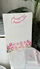 A beautifully styled wedding invitation setup is showcased in this elegant display. The main card features Arabic calligraphy reading 'مُحَرِّم' with the date '19.06.2026' and 'قِيلَتْ بِدُرُوْحَا تَشَارِي شُعُورِي', all set against a soft pink floral border with tulips and cosmos. Accompanying the main invitation are matching envelope details, including a pink wax seal and decorative bow. The scene is styled with marble surfaces and tropical greenery, creating a sophisticated, romantic atmosphere perfect for wedding planning inspiration. by @r200an with original sound