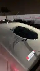 A close-up investigation of a maroon car with significant damage, including a large hole in the roof and a shattered rear window. The vehicle features 'SAUDI NATIONAL DAY' decals, suggesting a connection to the event. The scene is set at night, with people visible in the background near a wall with Arabic text. by @mo_kbs8 with انتهت القصه by 😂