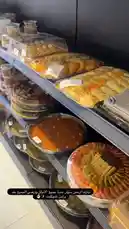 A close-up view of various traditional Arabic pastries and desserts arranged neatly on shelves inside a bakery. The display includes colorful, layered sweets, round trays of nutty confections, and other sweet treats typical of Middle Eastern cuisine. Arabic text is visible on screen, identifying the establishment as 'Brown Chocolate' and mentioning 'الإدراة وترفيس الجميع عند' (management and everyone's service). The scene suggests a focus on local, artisanal baking and customer service. by @ch11.30 with Original Sound by ch11.30