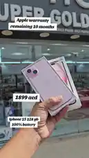 A hand holds a brand-new pink iPhone 15 in its original box, showcasing it in front of the 'Super Golden Mobile' store in Dubai. On-screen text highlights key selling points: remaining Apple warranty of 10 months, a price of 1899 AED, and a 100% battery condition. The phone is displayed outdoors in a sunny urban setting, emphasizing its excellent condition and value. by @super_golden7 with Original Sound by super_golden7