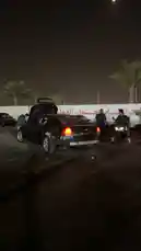 In a dimly lit parking lot at night, a heavily modified dark sedan with a custom rear bumper and open trunk is the center of attention. Several people gather around, including one on a motorcycle, creating a casual street car meet vibe. The car's engine roars aggressively, revving loudly as the scene unfolds. by @mo_kbs8 with Original Sound by mo_kbs8