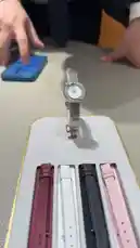 A close-up tutorial showing how to change a watch strap. A silver mesh bracelet watch is displayed on a stand next to four different colored leather straps—burgundy, white, black, and pink. A person's hands are seen interacting with the watch and a blue smartphone, demonstrating the process of swapping out the band. by @ab.7955 with Original Sound by ab.7955