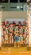 A group of young athletes, dressed in matching black and white striped jerseys, gather on a stage to celebrate their victory. They proudly hold gold medals around their necks and pose with multiple trophies, including a large central cup. Sparklers burn brightly on either side of the stage, creating a festive atmosphere. In the background, a banner displays Arabic text and logos, suggesting a formal sports event or tournament organized by a youth association. The mood is joyful and triumphant as the team celebrates their hard-earned success. by @playerassoc with original sound