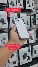 Watch as a hand holds and displays a white iPhone 15 with 128 GB storage. On-screen text highlights '87% battery' and 'iPhone 15 128 gb 1499 aed'. The background features shelves stocked with car phone holder boxes, suggesting a retail setting like محل السوبر الذهبي للهواتف. This video serves as a quick product showcase. by @super_golden7 with Original Sound by Contains: Koi Mil Gaya - Jatin-Lalit;Kavita Krishnamurthy;Udit Narayan;Alka Yagnik