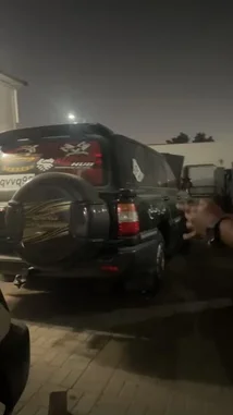 In this nighttime scene, a custom black SUV, likely owned by Ammar Sheikh, is the center of attention. The vehicle features a spare tire cover with 'Landrover' branding and a red graphic decal on the rear window. A person is seen filming the SUV with a smartphone, capturing its details under artificial lighting. The setting appears to be a parking area or a private lot, with the focus on the unique modifications and design elements of the SUV. by @raptor9431 with Original Sound by raptor9431